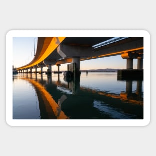 Morning sun strikes side Tauranga Harbour Bridge in golden hue reflected leading lines into calm water below Sticker
